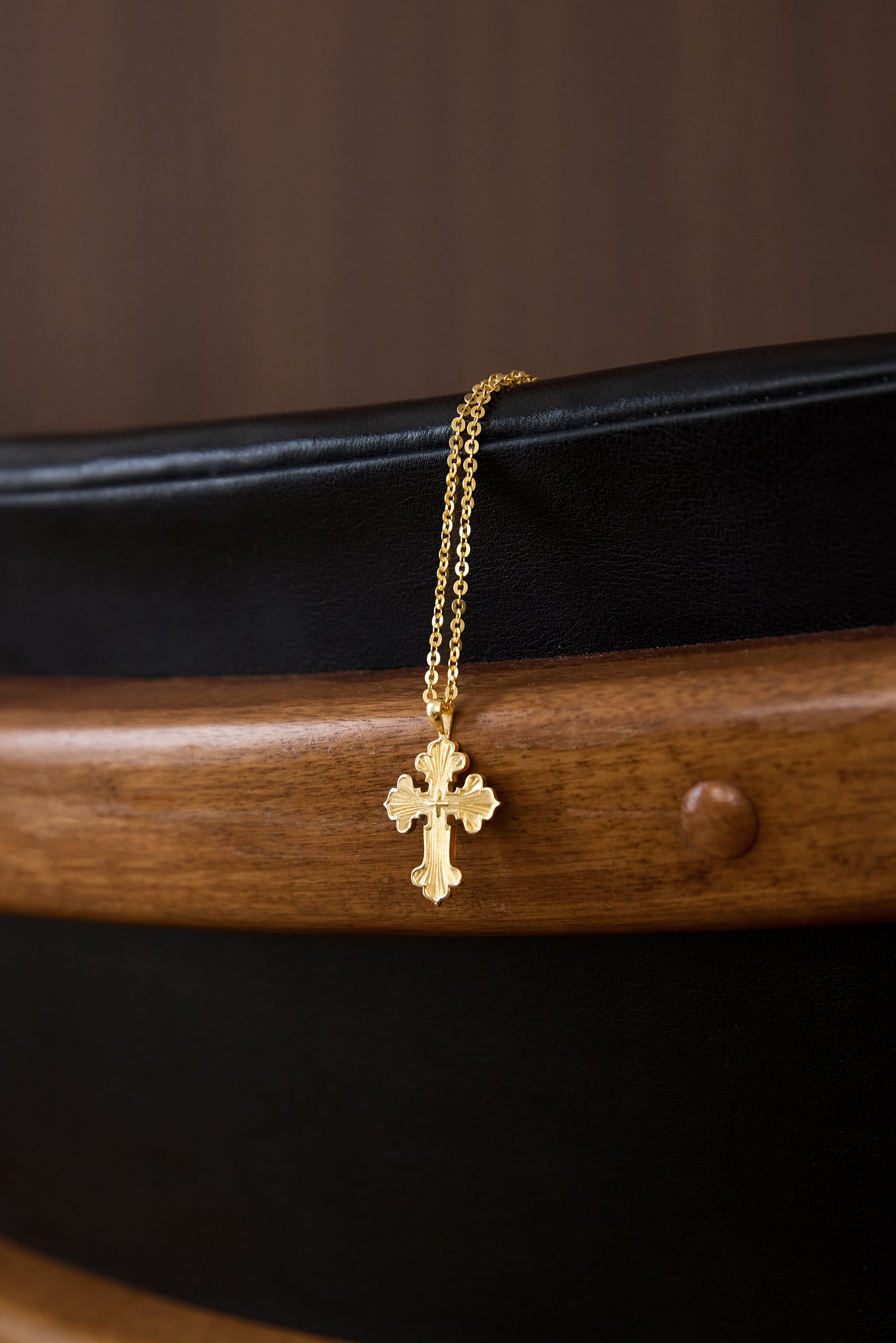 Tawalap cross pendant in 18k gold, handing on wooden chair.